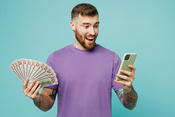 Young surprised fun man he wears purple t-shirt hold fan of cash money in dollar banknotes use...