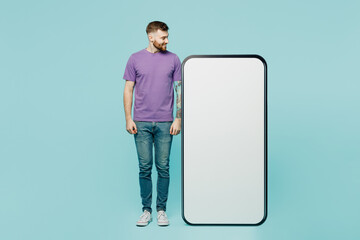 Full body smiling young man he wears purple t-shirt look at big huge blank screen mobile cell phone smartphone with area isolated on plain pastel light blue cyan background studio. Lifestyle concept.