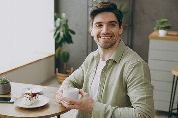 Side view young smiling cheerful happy caucasian man wear shirt casual clothes drink tea look aside sitting alone at table in coffee shop cafe restaurant indoors rest relax during free time inside.