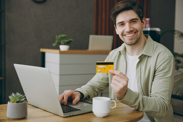 Young cheerful happy fun man wears casual clothes sits alone at table in coffee shop cafe indoors work using laptop pc computer hold credit bank card shopping online. Freelance mobile office concept.