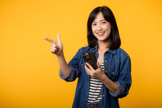 Portrait beautiful young asian woman happy smile dressed in denim jacket showing smartphone with pointing finger hand gesture to free space isolated on yellow studio background. app smartphone concept
