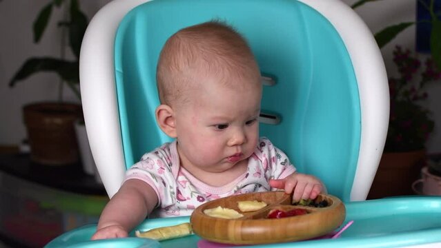 Cute baby girl learning how to feed herself and exploring food. Baby-led weaning (BLW) at 6-9 months old babies. Baby self-feeding in a high chair at home.