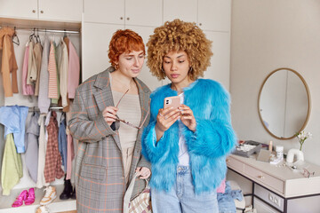 Two fashionable women concentrated attentively in smartphone chooses clothes in online store make...