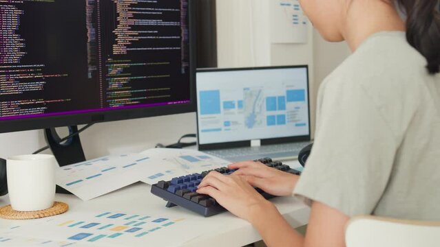 Close-up young Asia girl IT development programmer typing on keyboard coding programming fixing database code on computer monitor and laptop screen on table in workroom at house office.