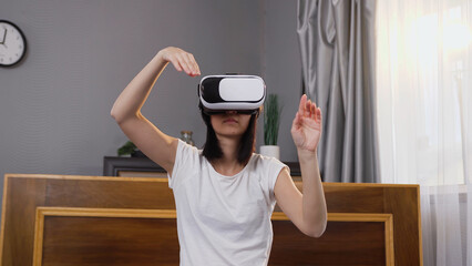 Beautiful concentrated young woman in virtual reality headset making scrolling on imaginary screen ,sitting on bed in contemporary bedroom