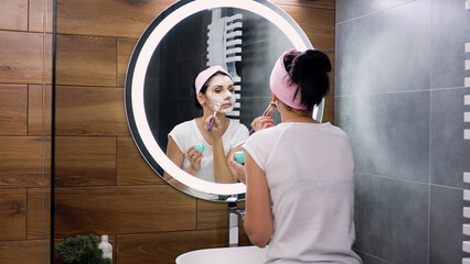 View from back where attractive concentrated young woman with headband cleaning her face with cosmetic foam, standing in front of mirror