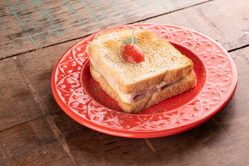 grilled ham and cheese sandwich on red ceramic plate decorated with cherry tomatoes and oregano on rustic wooden breakfast table in angled landscape