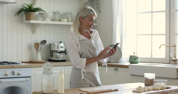 Happy chef blogger woman in apron baking in kitchen, taking picture of table with raw buns, flour, dough, using, Internet application on smartphone, smiling, turning look at camera