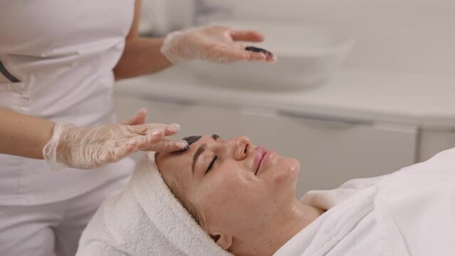 Beauty salon, Facial detoxification, Mud therapy. Beauty salon, cosmetologist administers black mask on client's face for facial detoxification, lifting, kelp rejuvenation, mud therapy