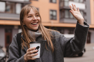 Smiling young woman waving her hand with a friendly cheerful smile to her neighbours. Girl wear grey sweater and coat, hold cup of tea, meeting friends, welcome, nice to meet you. Hi, hello gesture.  © zvkate