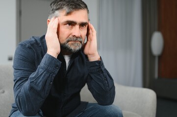Closeup of middle-aged bearded man suffering from headache at home, touching his temples, panorama with copy space. Migraine, headache, stress, tension problem, hangover concept