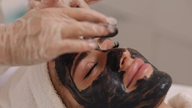 Kelp rejuvenation, Cleansing treatment, Mousse mask. Beauty salon cosmetologist puts black mask on patient's face, providing facial detox, lifting, algae rejuvenation, mud treatments, acne care