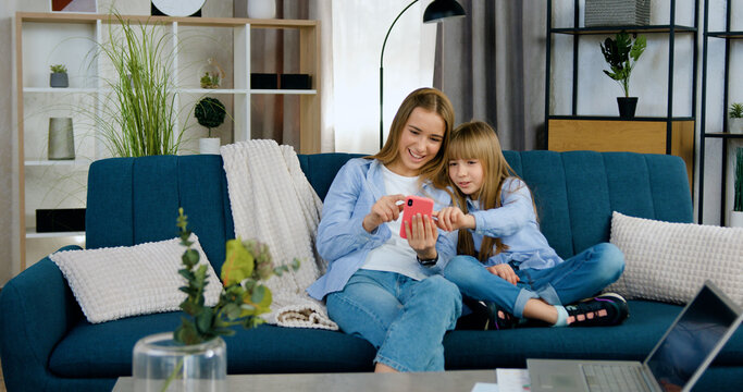 Adorable friendly carefree younger and eldest sister sitting together on soft couch in contemporary apartment and using smartphone to watch intersting videos