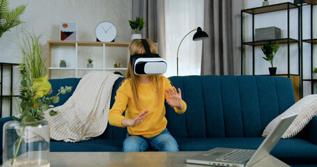 Beautiful interested satisfied 10-aged girl sitting on couch in living-room and gesturing on imaginary screen in augmented reality goggles