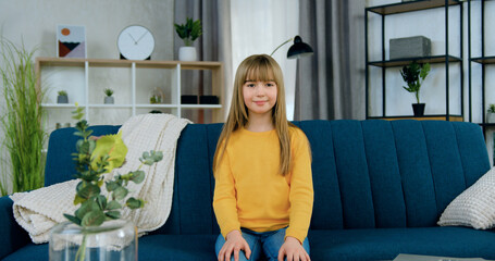 Beautiful smiling happy modern 10-aged long haired girl in casual clothes sitting on soft couch in...