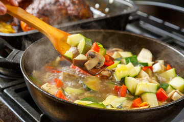 A frying pan with stewed vegetables and a wooden spoon on a stove next to a tray with baked chicken.