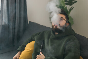 Bearded millennial or gen z man smoking hookah while relaxing on sofa at home - chill time and...
