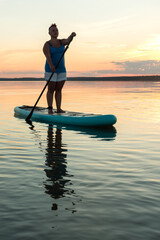 A woman in mohawk shorts stands on a SUP board with a paddle at sunset in a lake against the backdrop of a sunset sky and water.