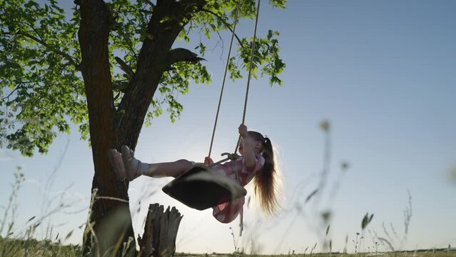 Concept of family happiness, dreams, holiday. Child swing, kid girl smile in flight. Child plays on wooden swing, dreams of flying. Happy little girl kid swings on swing in park under tree at sunset