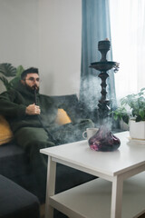 Bearded millennial or gen z man smoking hookah while relaxing on sofa at home - chill time and resting concept