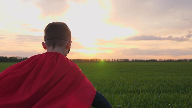 A happy boy runs in a red raincoat across a green meadow against the background of sunset. The silhouette of a child. The kid dreams of becoming a superhero.