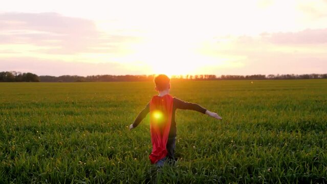 A happy boy runs in a red raincoat across a green meadow with his arms outstretched. against the background of sunset. The silhouette of a child. The kid dreams of becoming a superhero.