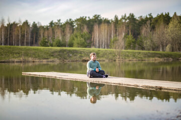 A boy is sitting on a bridge in a green park. The path is a bridge over the lake.