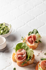 Bruschetta with ham or prosciutto, cream cheese, cucumber, spinach and microgreens. Tasty sandwich or toasted bread on white tile background. Tasty appetizer or breakfast, text space