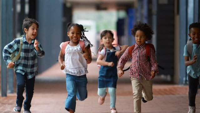 Handheld shot showing class of multi-cultural elementary school students running along outside walkway by school building - shot in slow motion