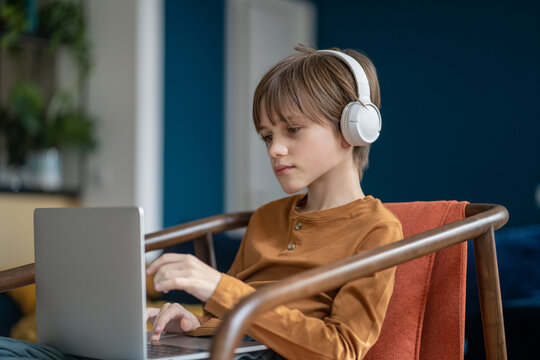 Serious attentive caucasian teen boy in headphones having online class on laptop sitting on chair in living room home. Remote, distance education, homeschooling. Listening lection, lesson, webinar.