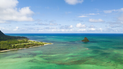 Hawaii's coastline with palm trees, ocean, clouds, and mountains in the background, aerial drone