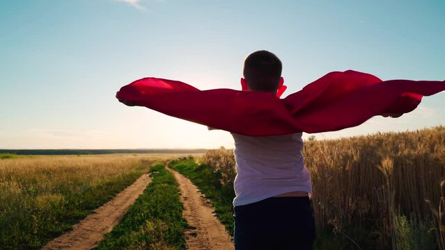 Superhero boy runs across green field, childhood dream to fly. Child, superhero in red cape, nature, sky. Active Boy, child plays superhero in sun. Happy run of child in red raincoat at sunset park.