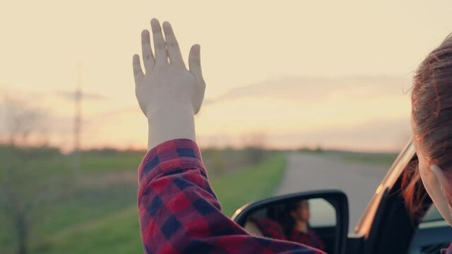 free woman travels by car catches the wind with her hand from car window. Girl with long hair is sitting in front seat of car, stretching her arm out window and catching glare of setting sun