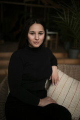 Beautiful woman in black sitting on sofa, looking at camera, room background.