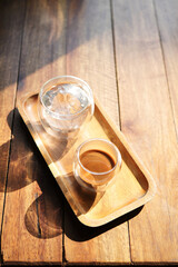 Hot coffee and glass of ice on wooden tray under sunlight