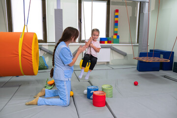 Child with physiotherapist on swing during sensory integration session. Little girl in...