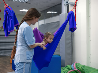 Physical therapist working with little girl in hammock made of elastic material. sensory...