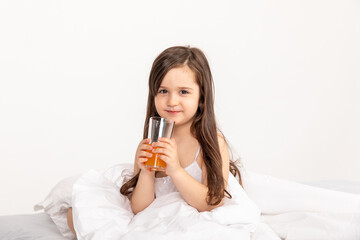 Happy little girl child in pajamas with a glass of fresh orange juice sitting at home in bed. Good morning