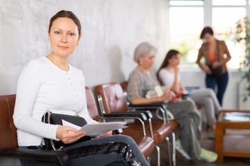 Fototapeta premium job female seeker is sitting in line with other women, holding paper documents in hands and carefully reading important information
