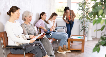 Adult woman in casual clothes reads text of document while sitting on chair in waiting room waiting for her turn