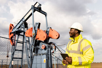 Oil field worker monitors drilling operations on oil rig with tablet and helmet.