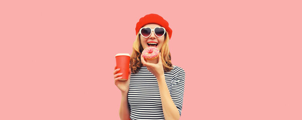Portrait of happy cheerful young french woman eating sweet donut with cup of coffee or juice in heart shaped sunglasses and beret hat on pink background