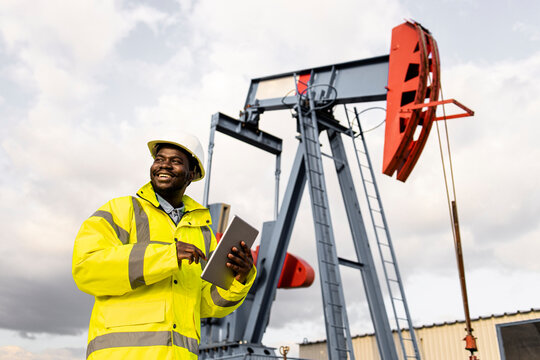 Oil field worker in protective work wear and helmet holding tablet by the oil rig.