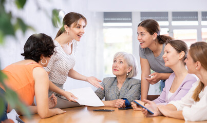 young woman sits on table, holds paper document in her hands and explains its essence to female colleagues present.Teamwork, concept of unconventional working atmosphere
