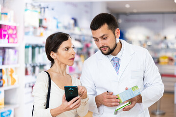 Fototapeta na wymiar Friendly man pharmacist helping young adult woman with electronic prescription in her phone choosing medical supplies at drugstore