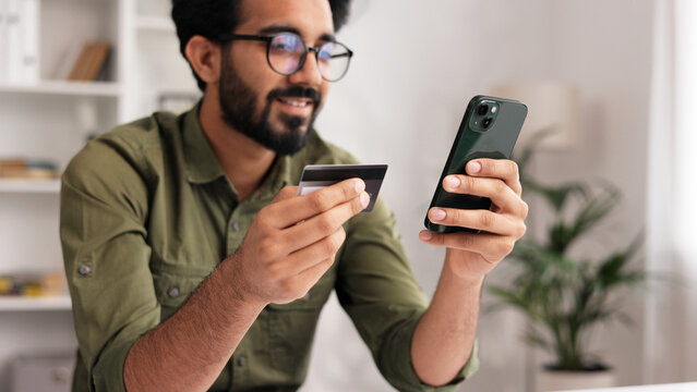 Smiling pretty young Indian brunette boy holding mobile phone and credit card sitting at the table. Happy man makes online shopping using mobile payment. E-banking app service.