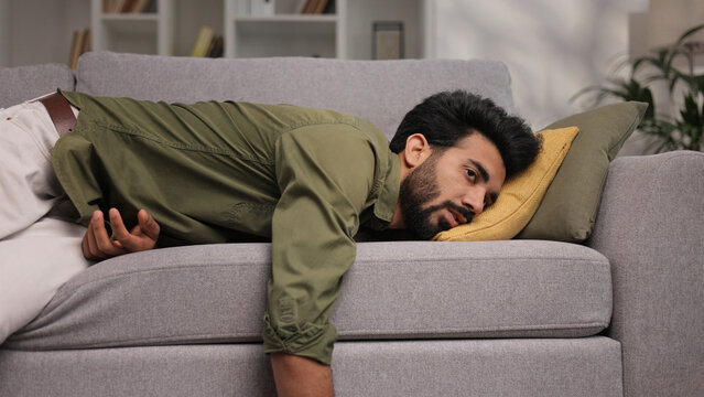 Tired Indian boy slumps on couch feeling listless. Feeling depressed, lack of motivation, sadness, boredom. Exhausted, he collapses on the couch from stress. Burn out.