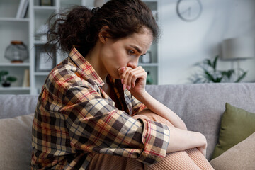 Young girl is deep in sad thoughts, thinks, looks away, feels tense and unhappy. Brunnete woman sitting on the couch have problen in personal life, feel depressed and broken.