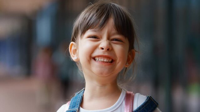 Head and shoulders portrait of smiling female elementary school student wearing backpack standing outside school building - shot in slow motion