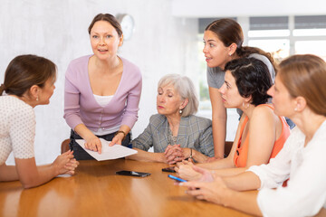 Smiling interested woman conducting informal work meeting with mixed age group of female colleagues sitting around table in office, discussing new strategies, projects, or work-related problems